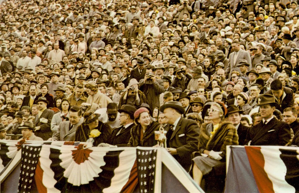 Color photo of Queen Elizabeth in the stands, with the crowd behind her.  