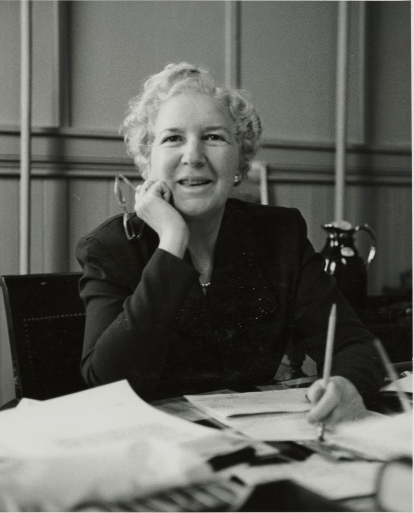 Adele Stamp sitting at her desk. She is in her middle years, with short light hair, glasses in one hand, and a blazer with a sparkly lapel. Her desk has many papers on it, and she is posed as if she were writing, but she is making eye contact with the camera.