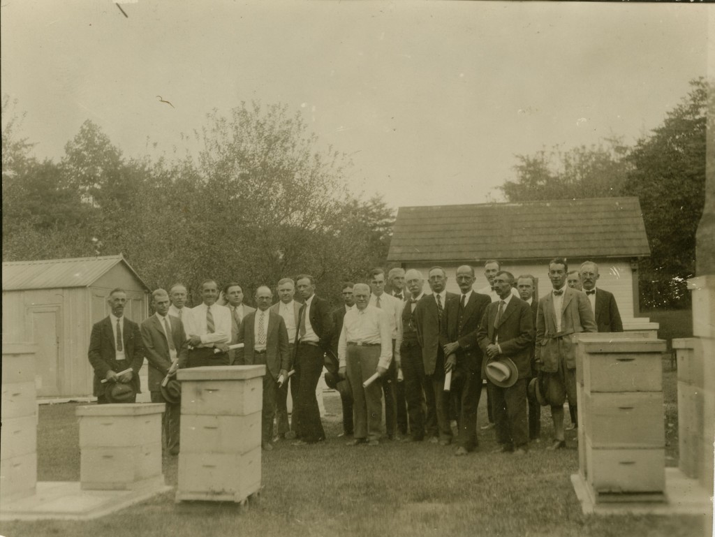 A large group of men stand outdoors in front of an orchard. There are also two small sheds behind them. In front of them are several beekeeping boxes. The men are dressed in suits, ties, and white collared shirts, and some of them hold hats in front of them.