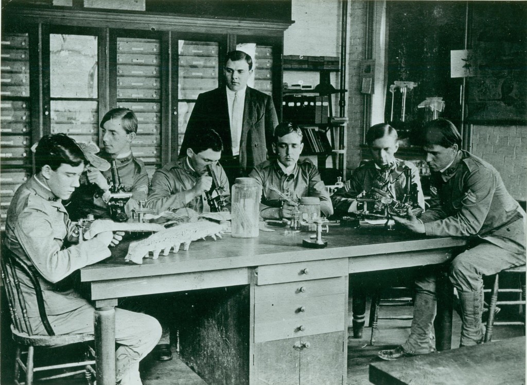 Six students sit around a wooden table. They are all wearing cadet uniforms and have side parts with their bangs swept to the side, and appear to be in their 20’s or early 30’s. The second student on the left is T.B. Symons. On the table are several insect models, microscopes, various scientific instruments, and a few glass jars. A professor in a dark suit and tie, believed to be Willis Johnson, stands over the table. In the background are a chalkboard, a small bookshelf with binders and small boxes on it, and a massive wooden cabinet with a dozens of specimen drawers.
