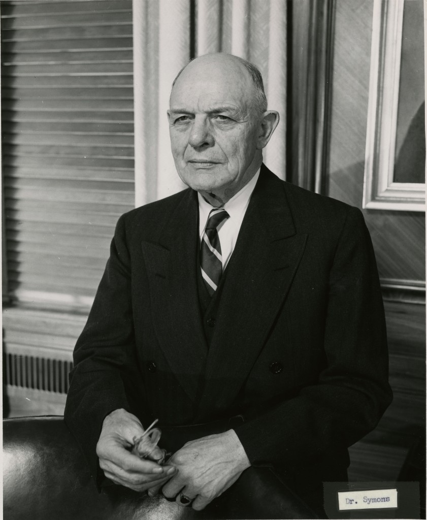 T.B. Symons stands in a nondescript office, resting his hands on the back of a leather chair. He is holding a pair of wire- rimmed glasses and is looking into the distance. He is bald and appears to be in his 60s or 70s, and is wearing a dark, double breasted suit and tie.