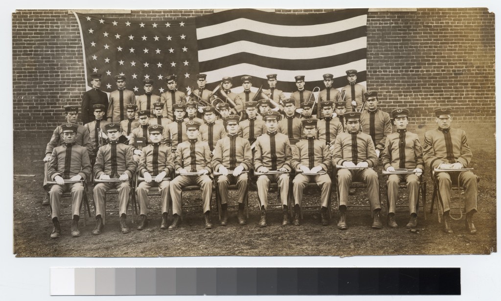 Four rows of young men in military uniforms, the first three rows of which are seated while the back row is standing. One of the men in the back row is older and in a darker uniform, presumably the officer who oversaw the band. Some of the young men are holding their instruments; the front row all have trumpets resting on their laps while the back row is standing with larger brass instruments. The middle rows are mostly obscured so it is unclear whether or not they have their instruments. Behind them is a massive American flag, which is pinned onto a brick wall.