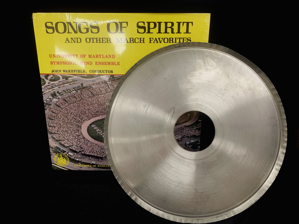 The vinyl album for ‘Songs of Spirit’ is propped up on a black background. The cover for the record is visible, but the vinyl record itself is not. There is a picture of Maryland Stadium on the front of the cover, and it is still wrapped in plastic, which reflects light. A large aluminum disc is propped up on it, partially obscuring the cover. 