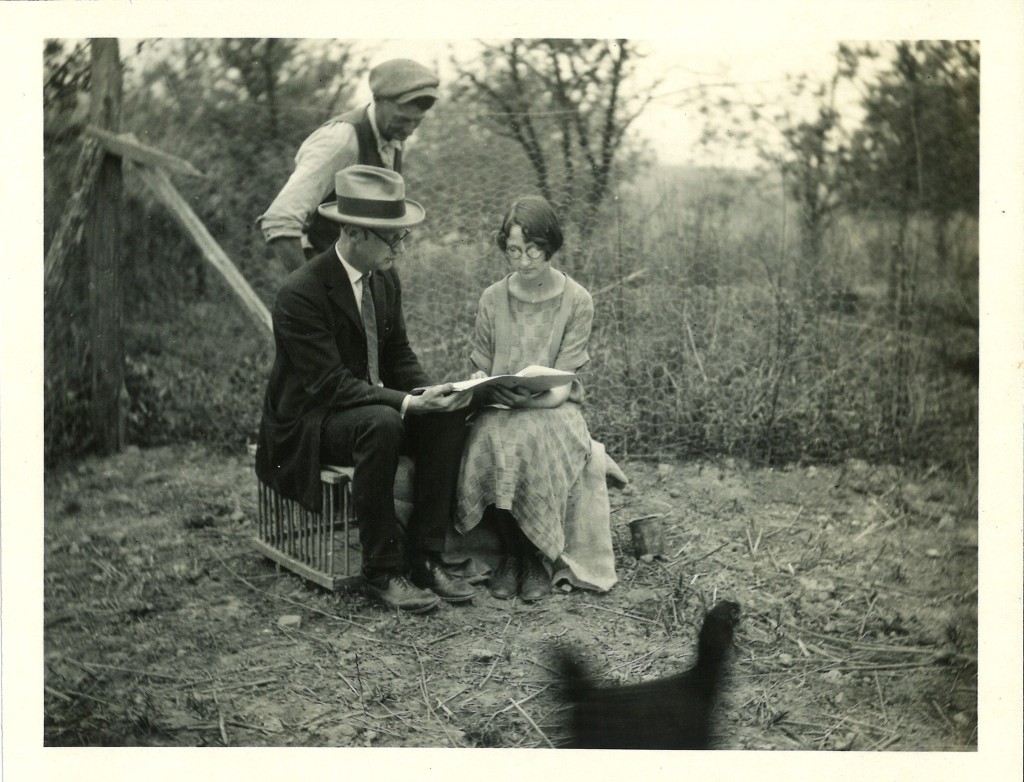 Three people are in frame, one young woman and two men in hads. One man and the woman, Edith Hobbs, are sitting on top of a wooden cage, presumably intended for chickens. They are each holding one side of a large book and appear to be reading it. The third man is standing over them, presumably also reading. There is a blurry black chicken running past the front of the frame. 