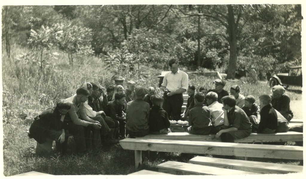 A group of around 20 young boys sitting on stumps and wooden benches. Their backs are facing the camera or they are only visible in profile, and an adult man in a white button-up shirt stands in front of them, holding eggs for some sort of demonstration.
