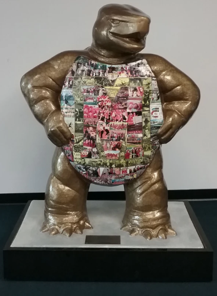 Color photo of a large statue of Testudo. She is standing upright on her back legs, with her front arms bent, hands resting on her waist. Her head, arms, and legs are golden. Her belly, where the shell is, is covered in numerous small photographs of students, faculty, staff, etc. of UMD at different events, and of different time periods. Some photos are color and some are black and white.