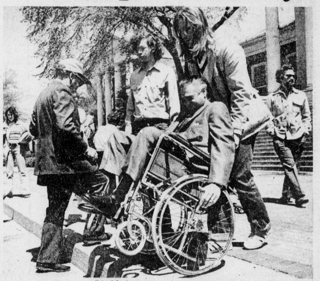 Black and white photo of student protesters. One man in a wheelchair is being carried down a large step. There is one man holding his wheelchair by its handles, by the man's head, and another man is at his feet, lifting the wheelchair's wheels up. Behind them, students walk past. One student is standing beside the group looking out.
