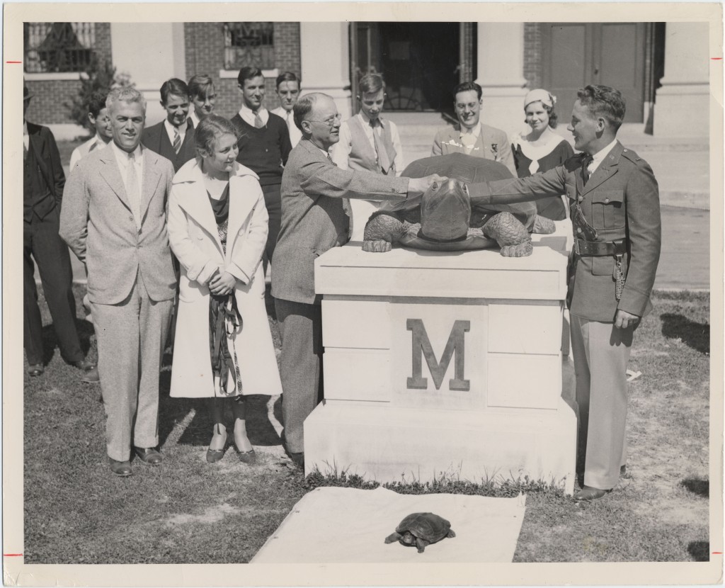 Black and white photo of the Testudo statue unveiling ceremony. In front of a building on a grass field is a large statue of Testudo, sitting on a square, White pedestal with a large letter "M" in the middle. To the left of the statue are three people standing and one person is standing to the right of the statue. 