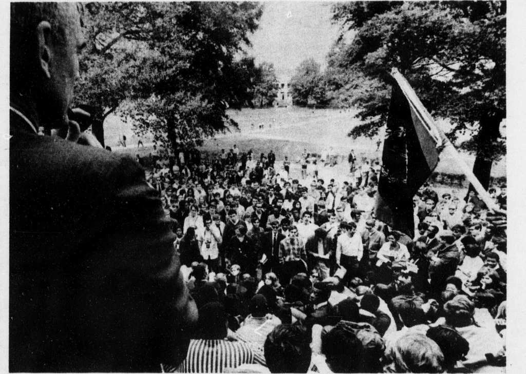 A black and white image of the crowd of students gathered outside the Home Economics building to protest. There are trees and flags in the background, and a man smoking a pipe blocking the camera's view of the left side of the crowd. 