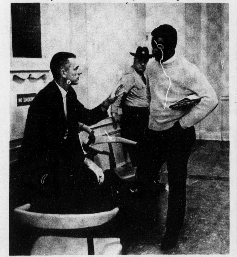 A black and white newsprint photograph of Bob McLeod, standing on the right, talking to a seated J. Winston Martin inside Marie Mount Hall. A state trooper in uniform stands in the background.