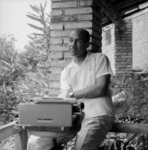 A black and white image of Ralph Ellison sitting behind a type writer on his porch. He is wearing dress pants and a white shirt, and gazing off to the side. 