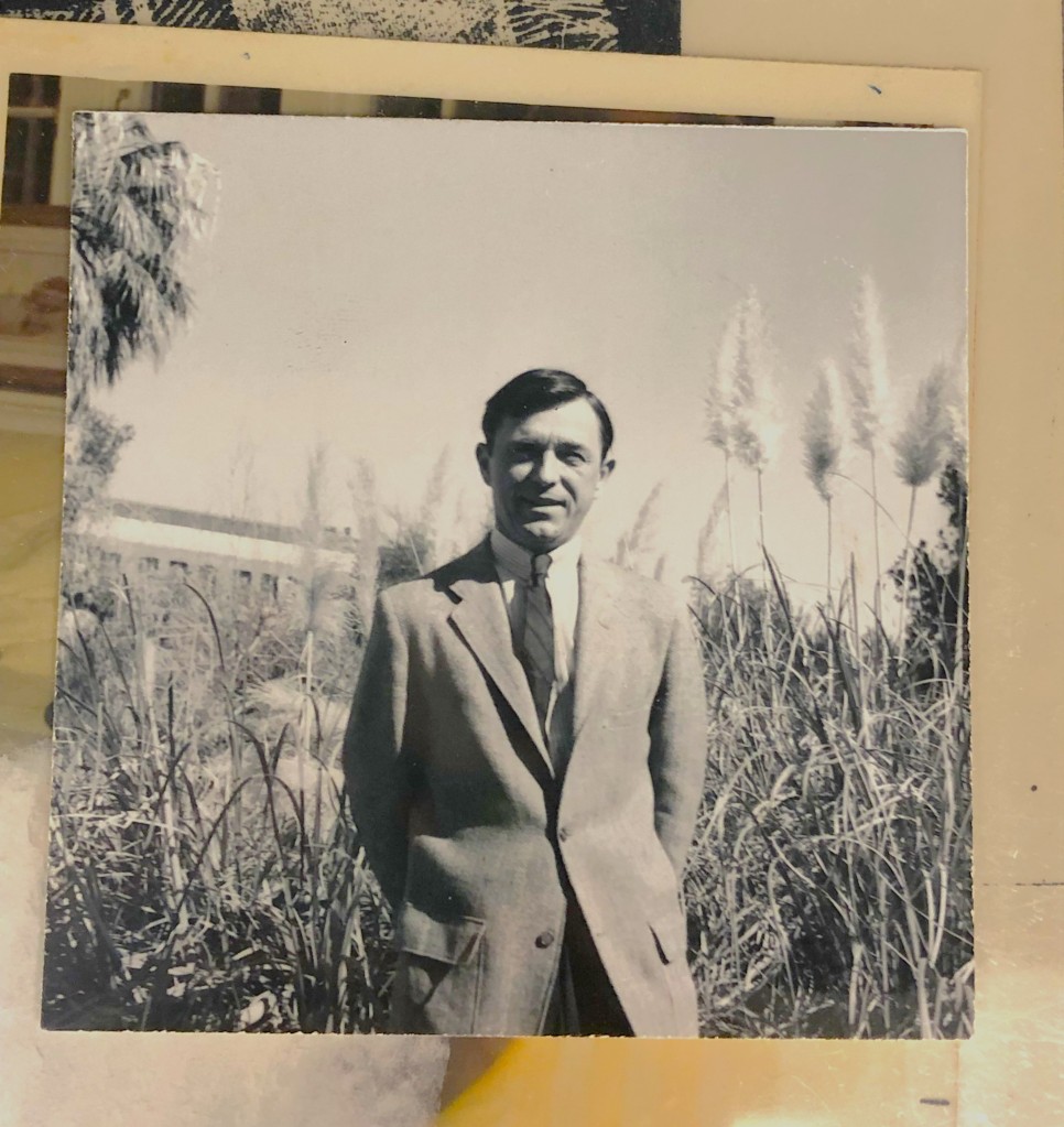Black and white photo of Whittemore standing in front of tall grass-like plants. He is wearing a suit and smiling directly at the camera. 